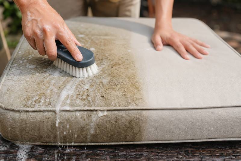 Outdoor Cushion Cleaning Tips: Close-up of a hand gently scrubbing an outdoor cushion with a brush and foamy solution, demonstrating effective outdoor cushion cleaning tips.